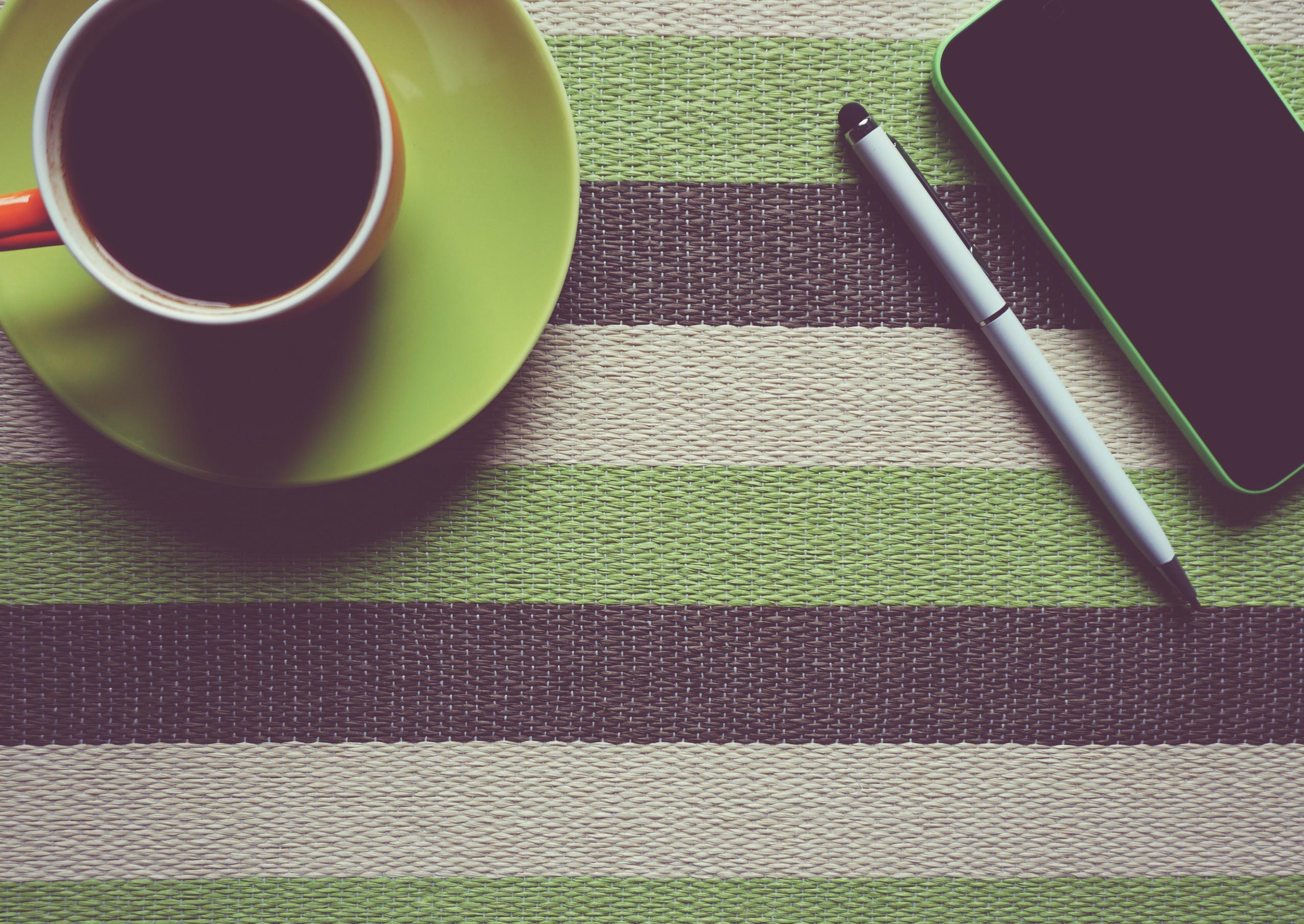 A cup of coffee on a table next to a cell phone and a pen.